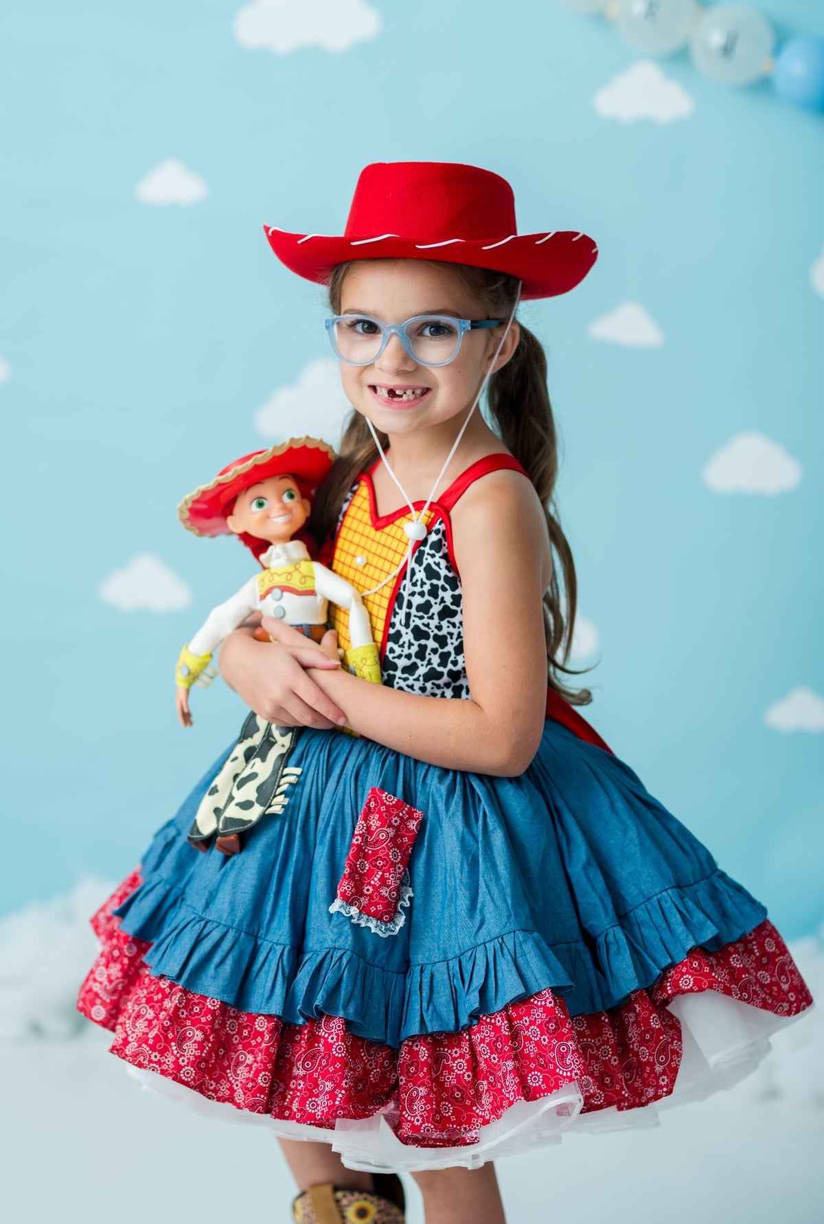 A young girl wearing a red cowboy hat and a blue and red ruffled dress with a cow-print bodice, holding a Woody doll against a light blue cloud-patterned backdrop.