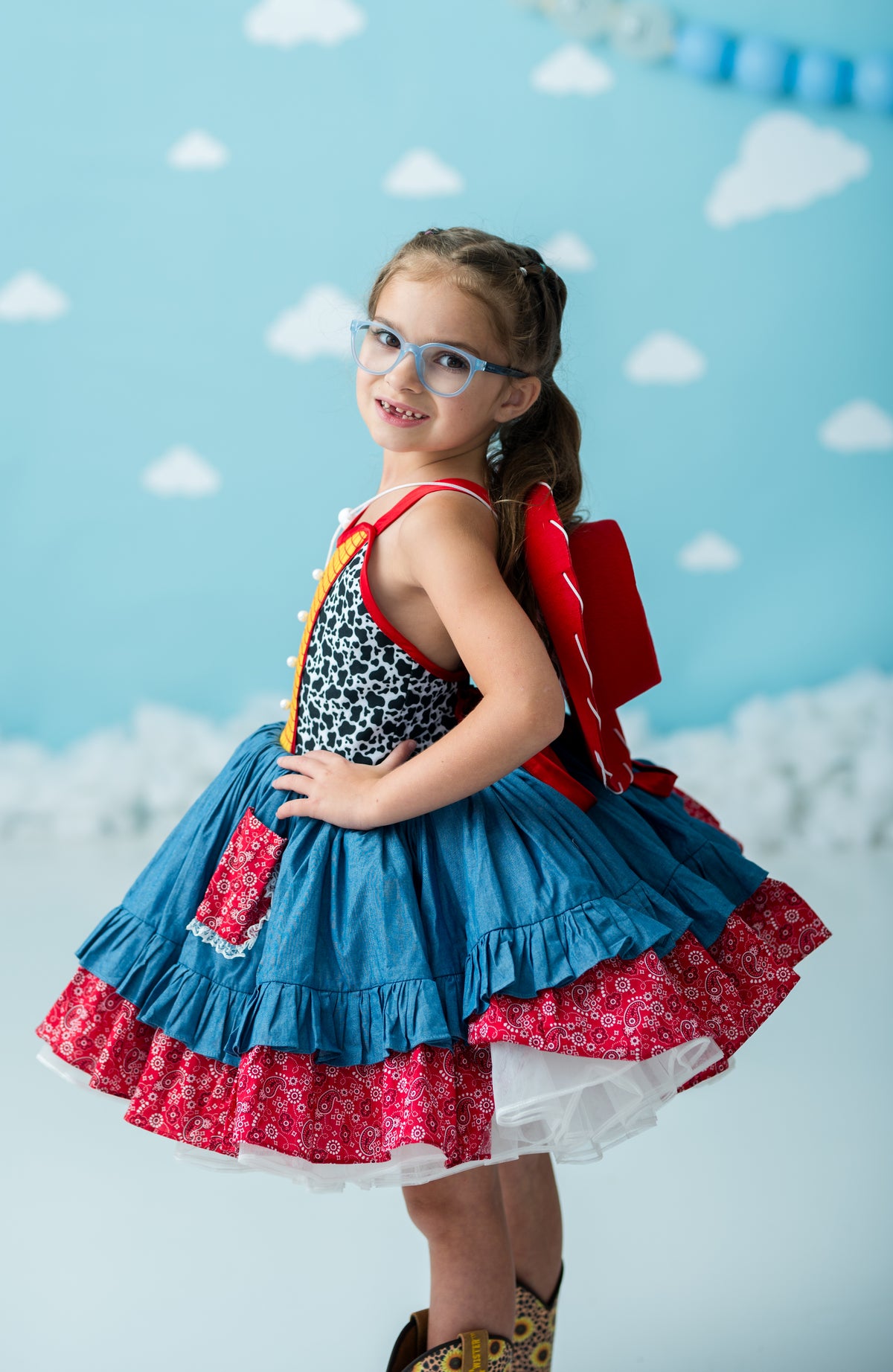 A young girl modeling a whimsical costume dress with a blue ruffled skirt, red patterned underskirt, black-and-white leopard print bodice, and red wings against a light blue cloud-patterned backdrop.