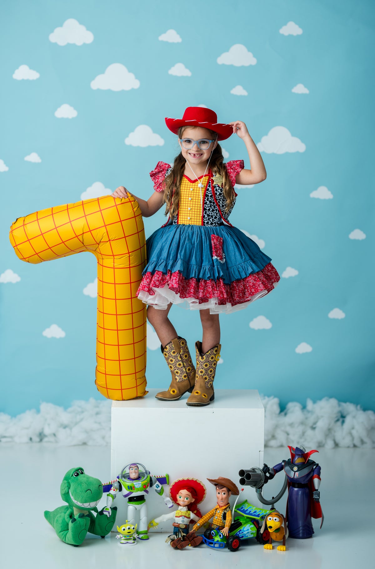 A young girl in a Woody-inspired cowboy costume stands on a white pedestal, holding a large yellow number 7 prop against a blue sky with clouds backdrop. Toy characters are lined up at the bottom of the pedestal.