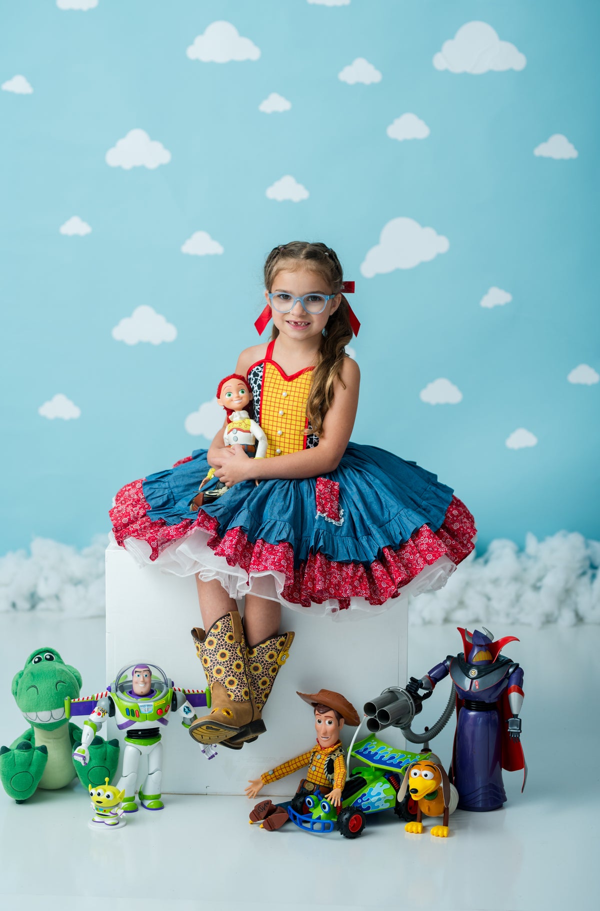 A young girl with braided hair and glasses sits on a white pedestal, wearing a colorful ruffled costume with a yellow top, blue skirt, and red trim. She holds a doll, with various toy characters arranged around her on a light blue cloud-patterned backdrop.