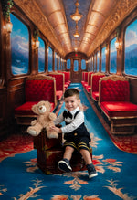 Child in formal attire sitting on a plush train seat with a teddy bear, surrounded by red seats and ornate decor.