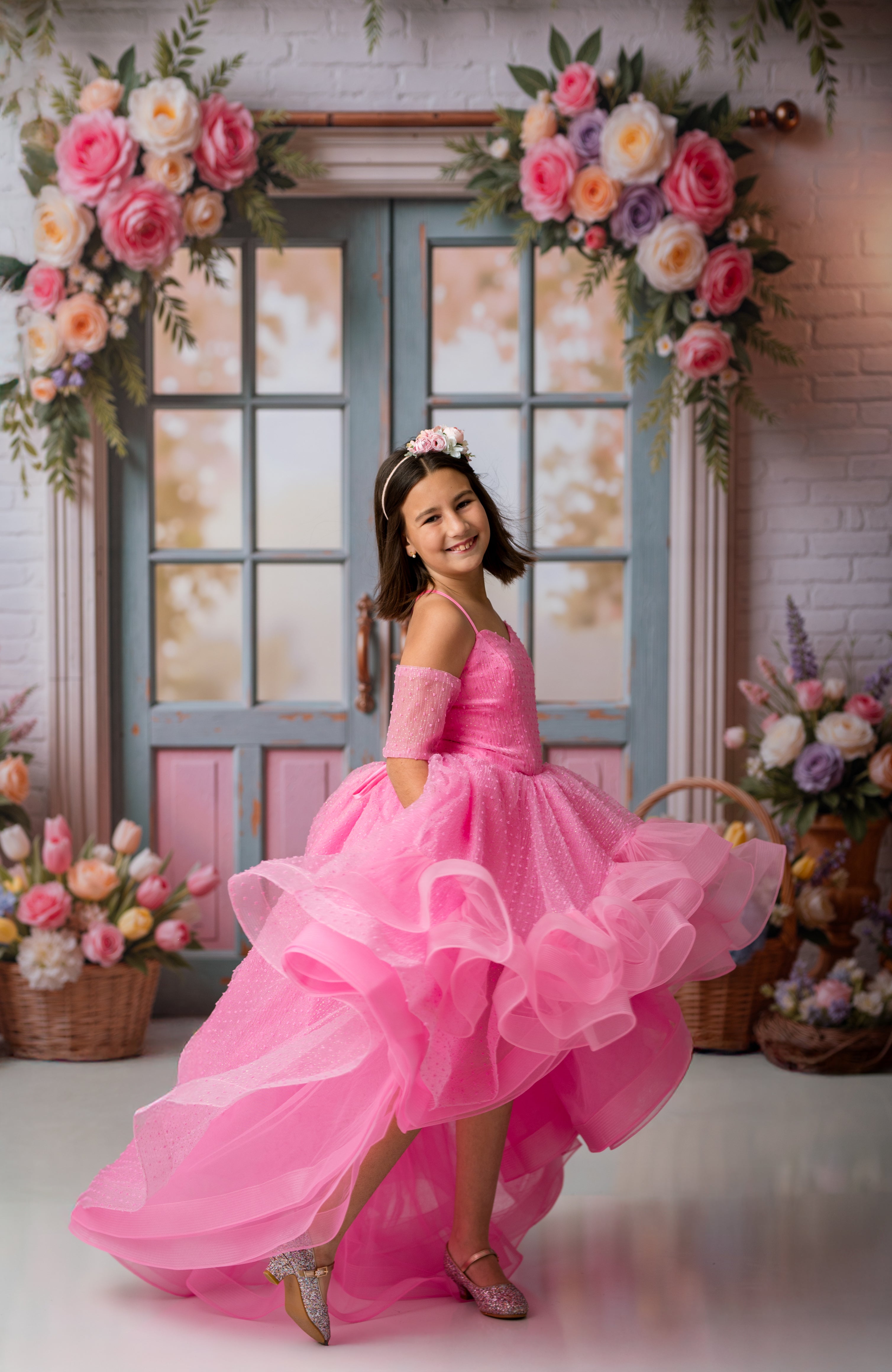 Young girl in a pink dress standing in front of floral decorations and a window.