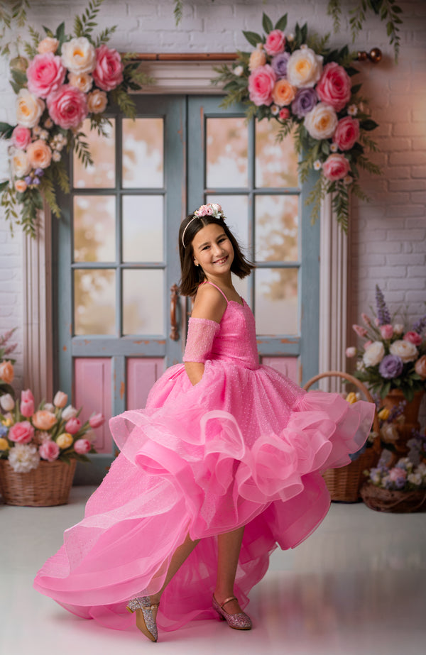 Young girl in a pink dress standing in front of floral decorations and a window.