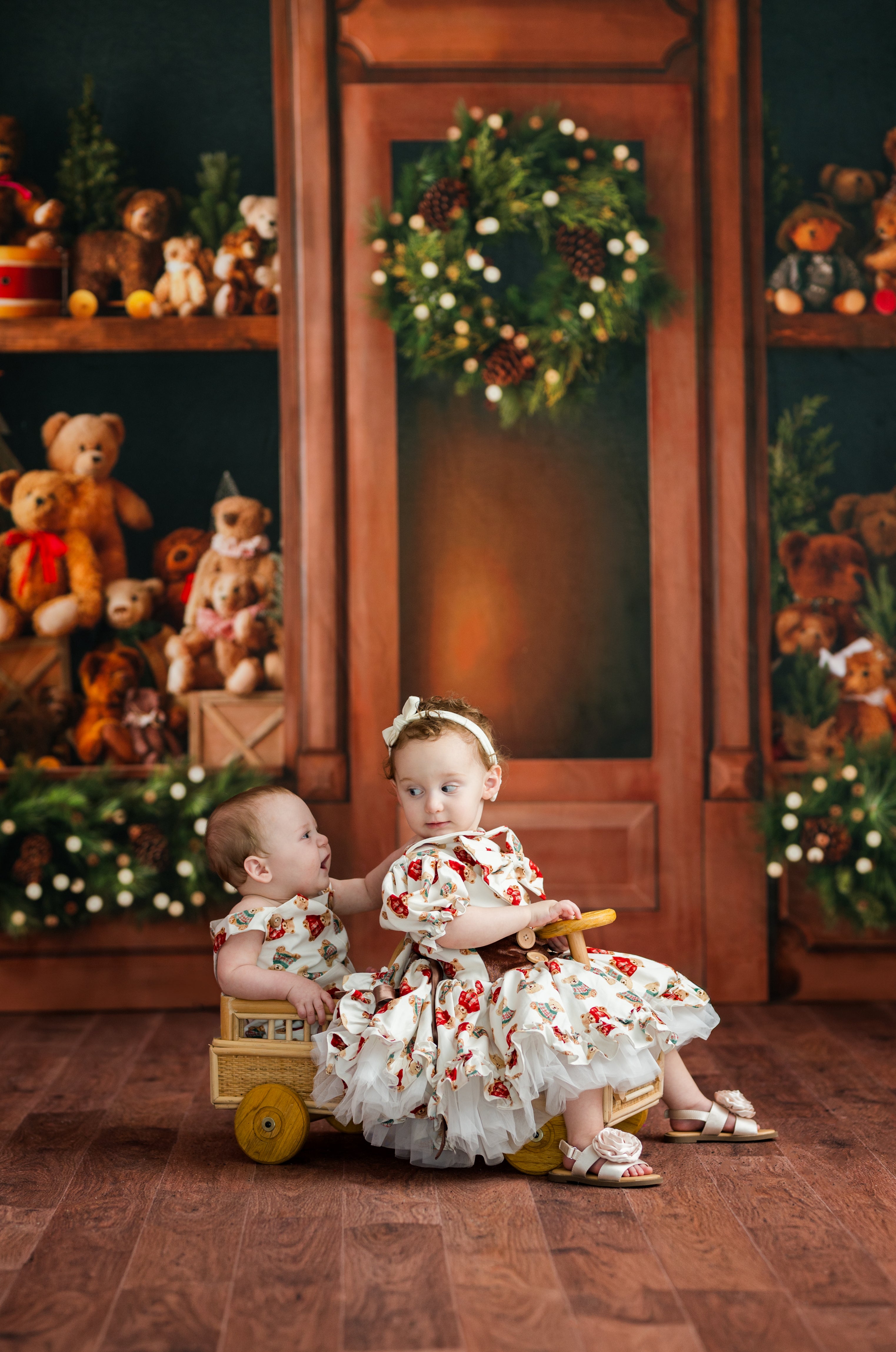 Two young children in matching white dresses with red and green floral prints pose with a small wooden toy cart in a decorated holiday-themed studio setting with teddy bears and wreaths.