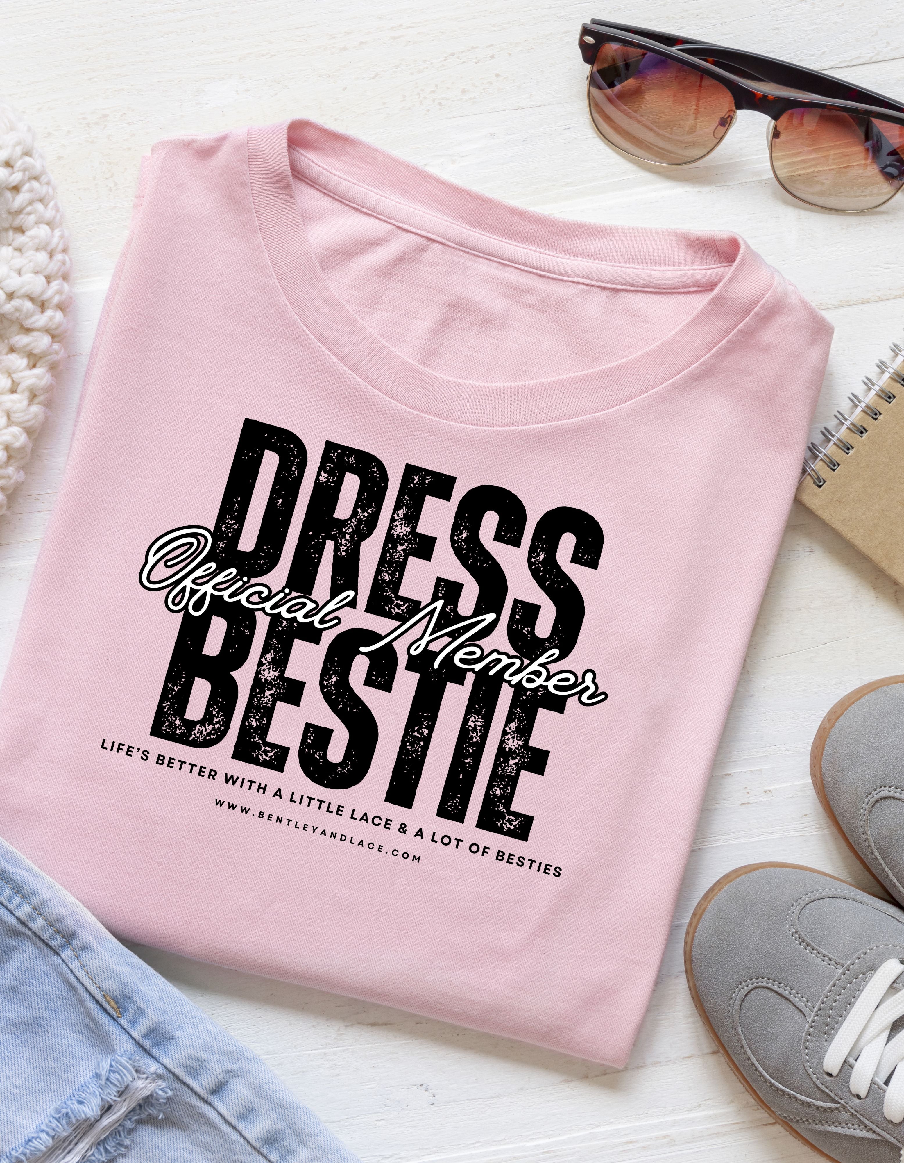 Pink t-shirt with 'Dress Bestie' text on a white surface with accessories.