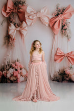 Young girl in a pink dress standing in front of a decorative wall with large pink bows and flowers.