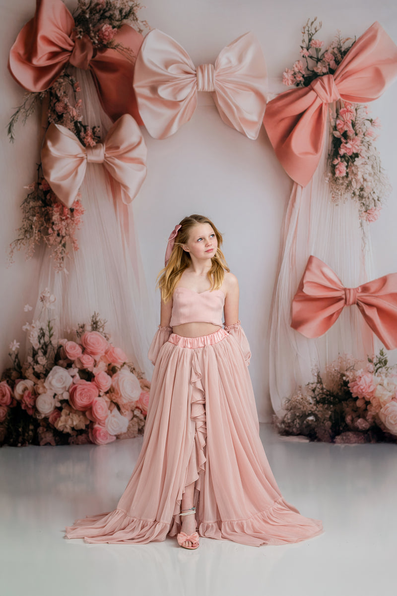Young girl in a pink dress standing in front of a decorative wall with large pink bows and flowers.