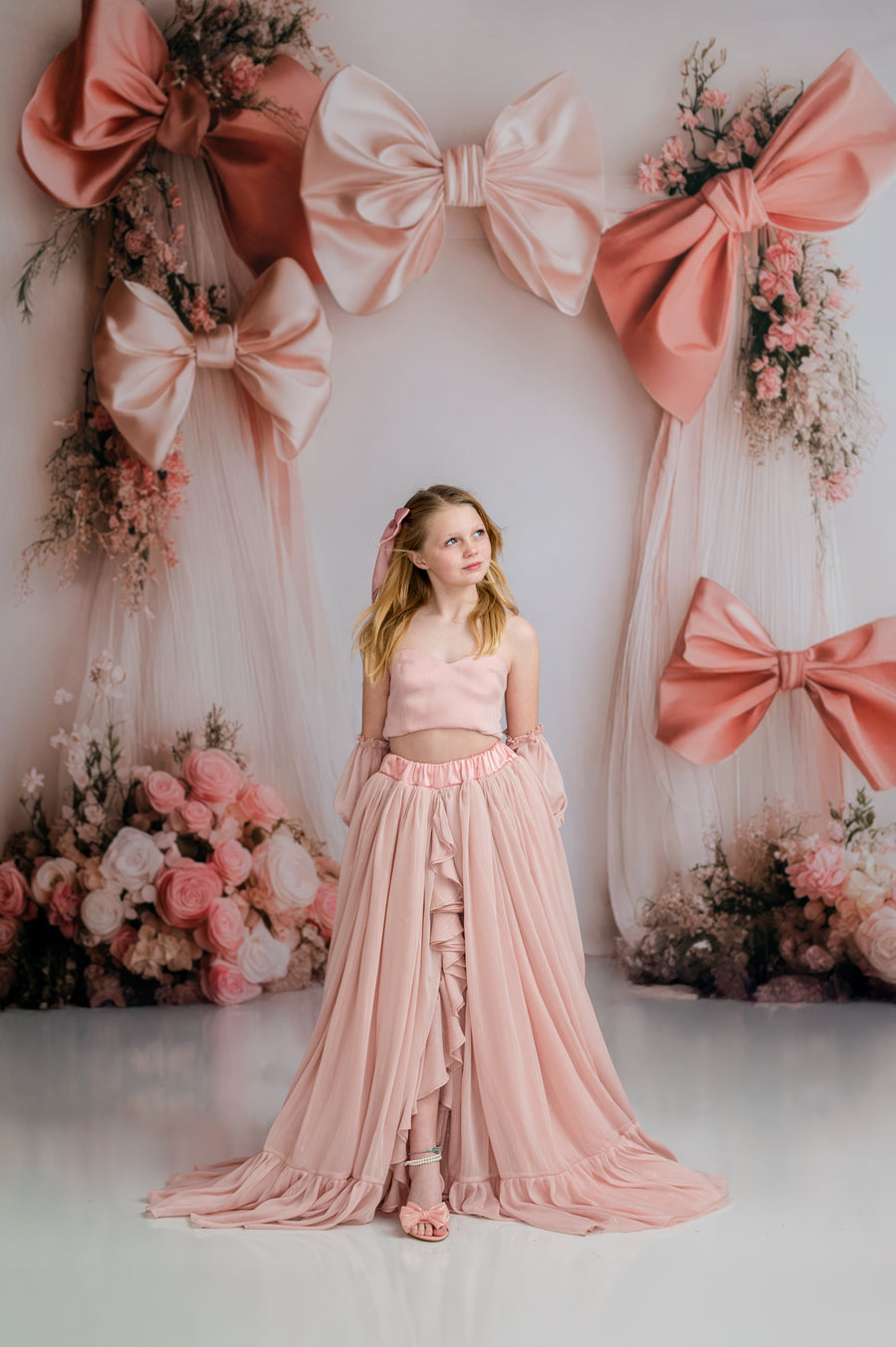 Young girl in a pink dress standing in front of a decorative wall with large pink bows and flowers.