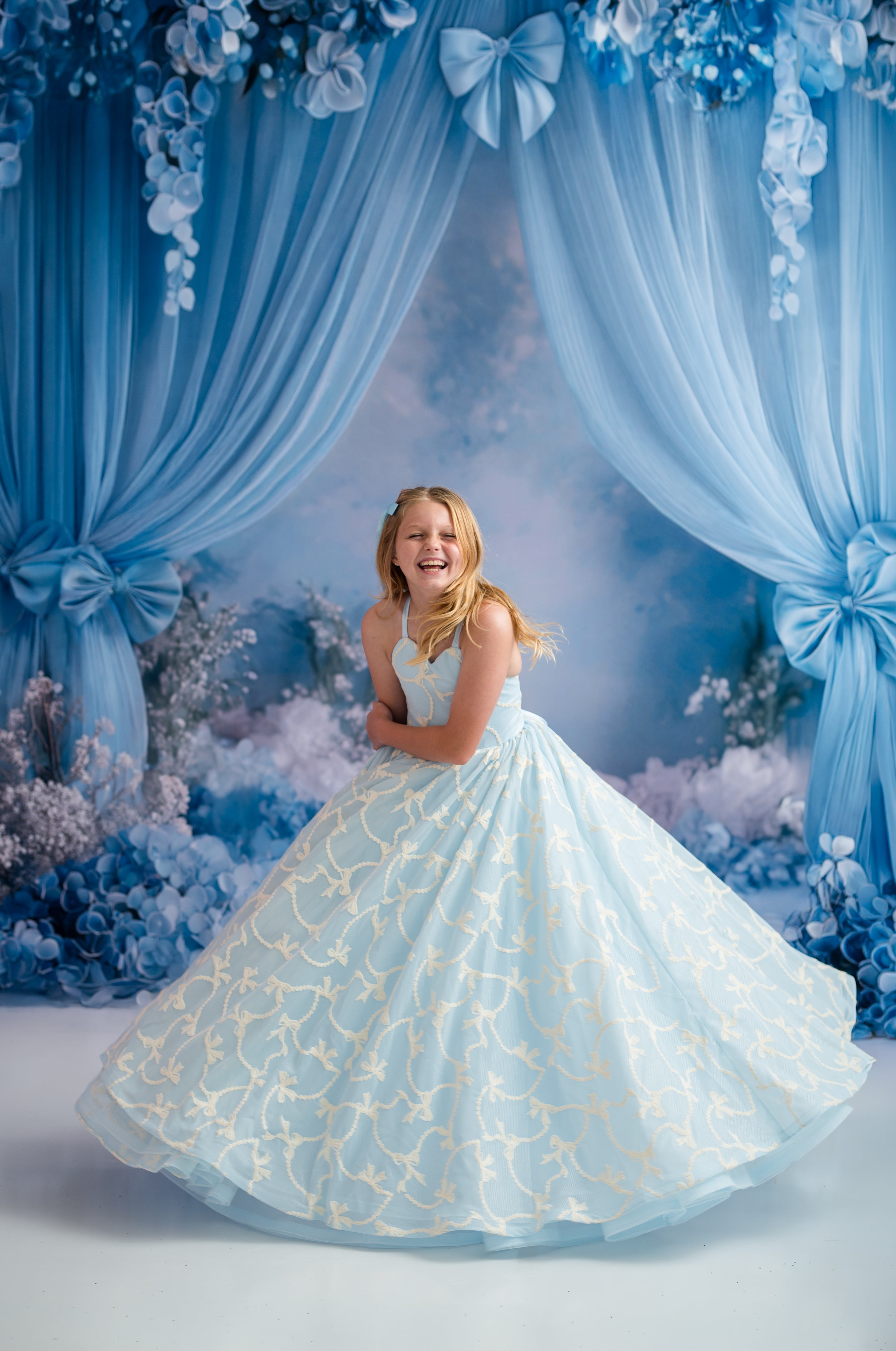 Young girl in a light blue dress with floral patterns standing in front of a decorative backdrop with blue curtains and flowers.
