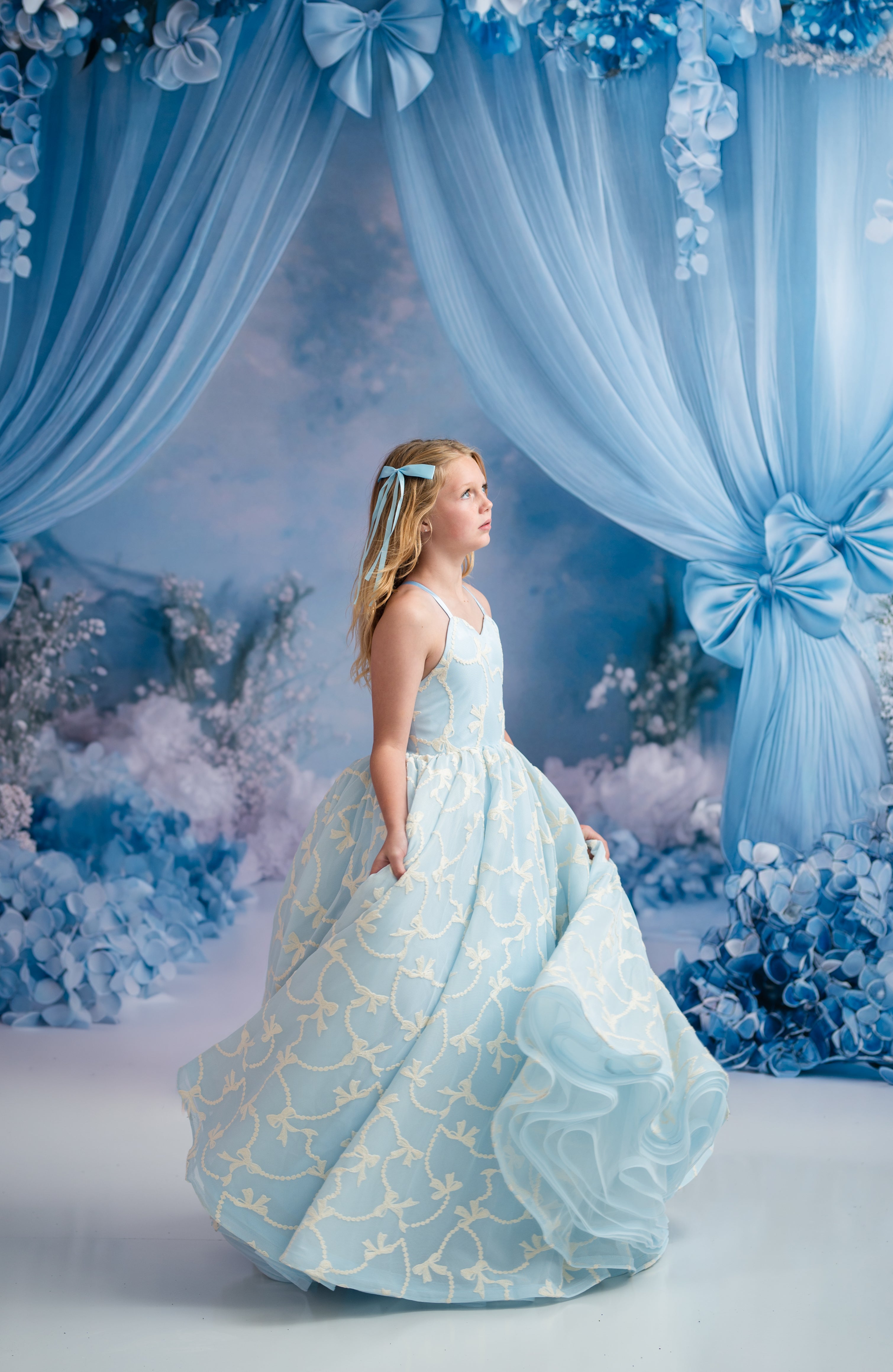 Young girl in a light blue gown with gold patterns standing in front of a decorative blue curtain and floral backdrop.