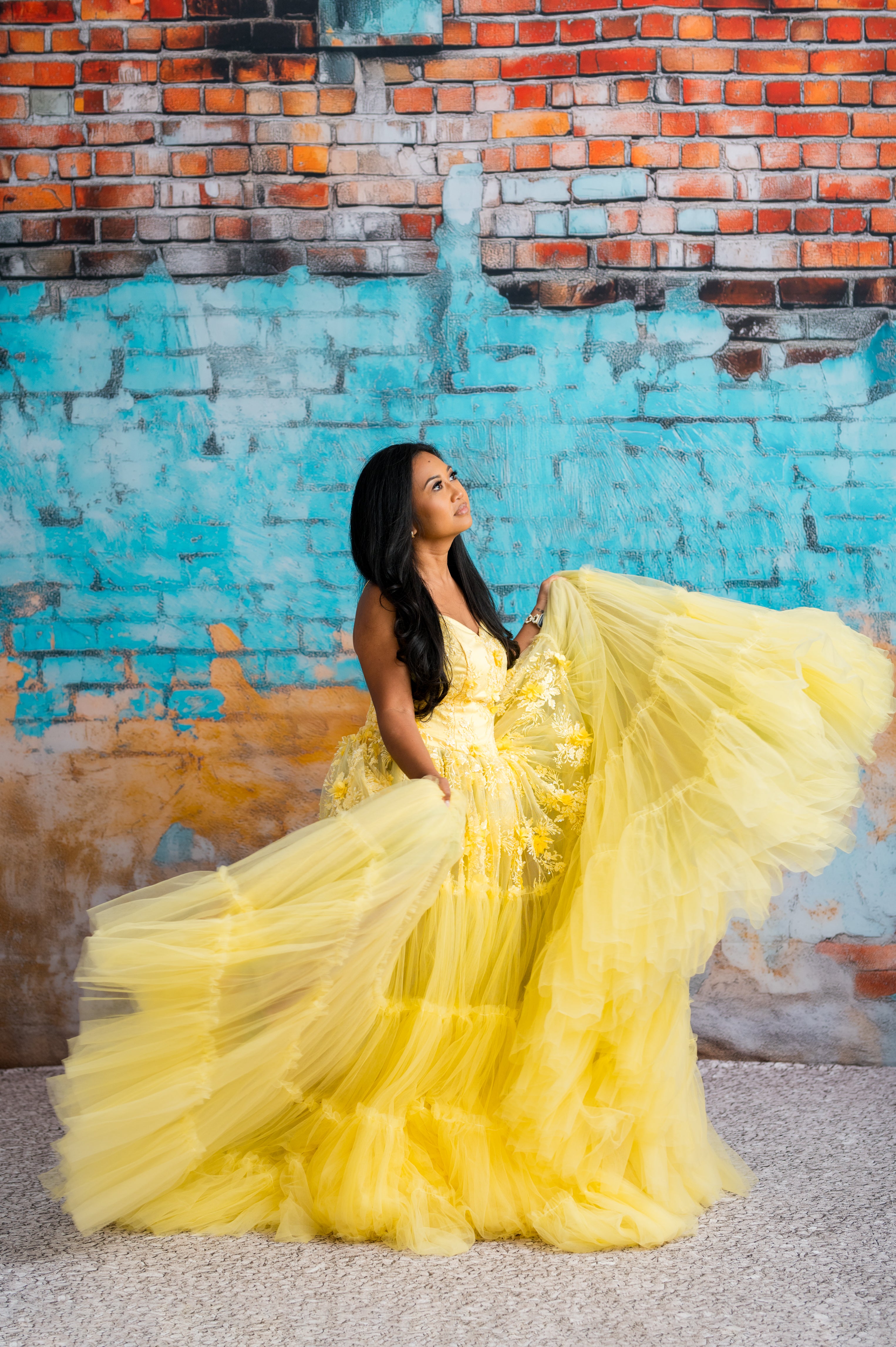 Woman in a yellow dress standing against a colorful brick wall.