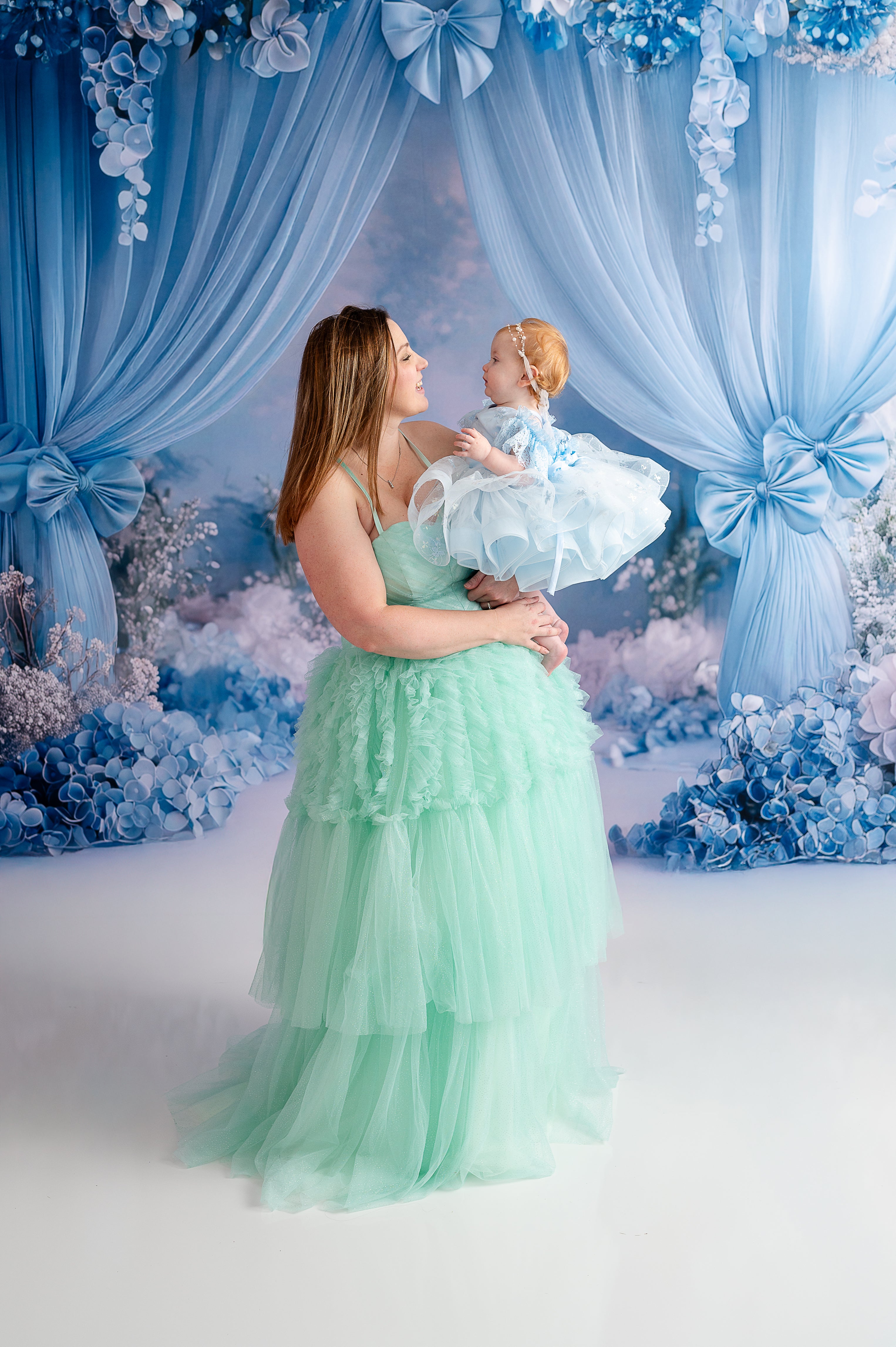 A young girl in a voluminous teal and gold gown looks up at her mother, who wears a matching mint green ruffled dress.
