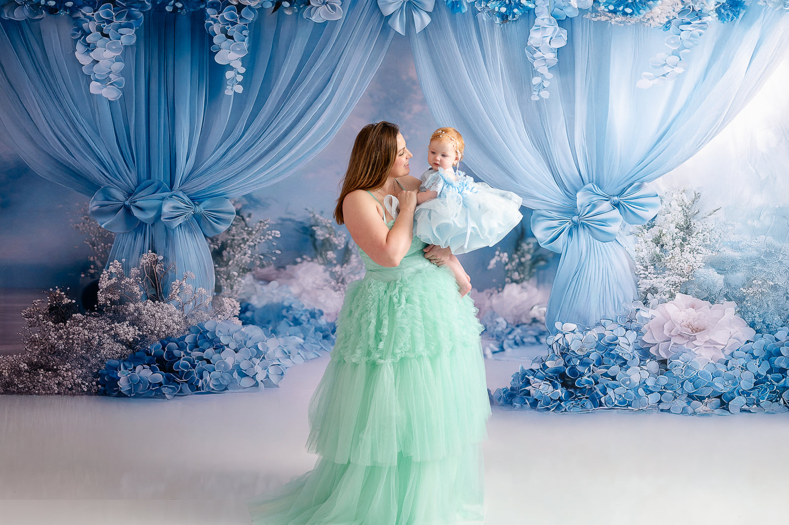 A mother in a mint-green ball gown stands gracefully beside her daughter, who wears a matching princess-style dress.
