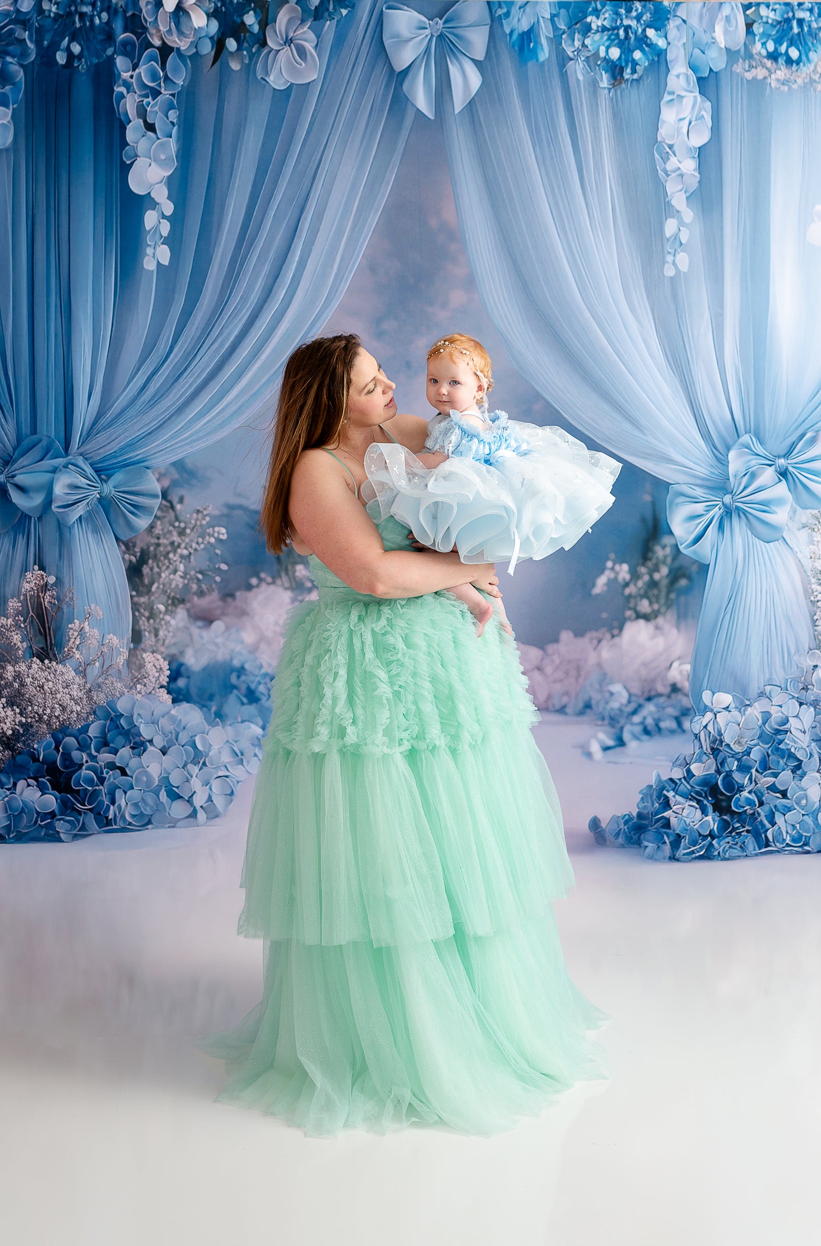 A woman in a flowing mint green gown gazes lovingly at her young daughter, who wears a coordinating teal and gold dress.
