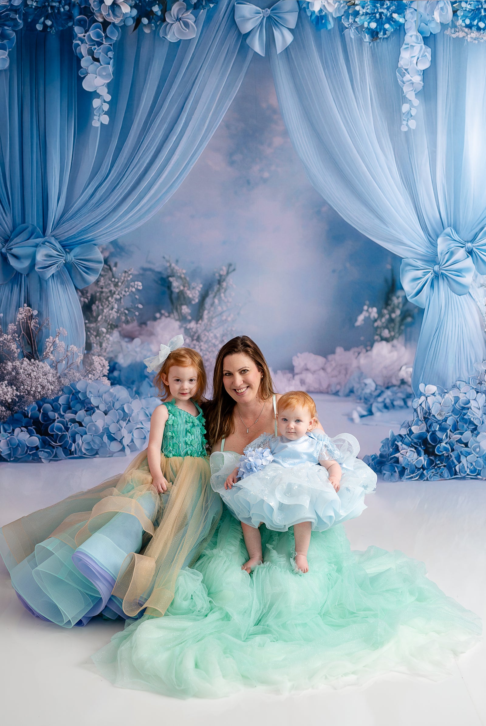 A whimsical mother-daughter portrait featuring layered pastel dresses and a soft floral backdrop.
