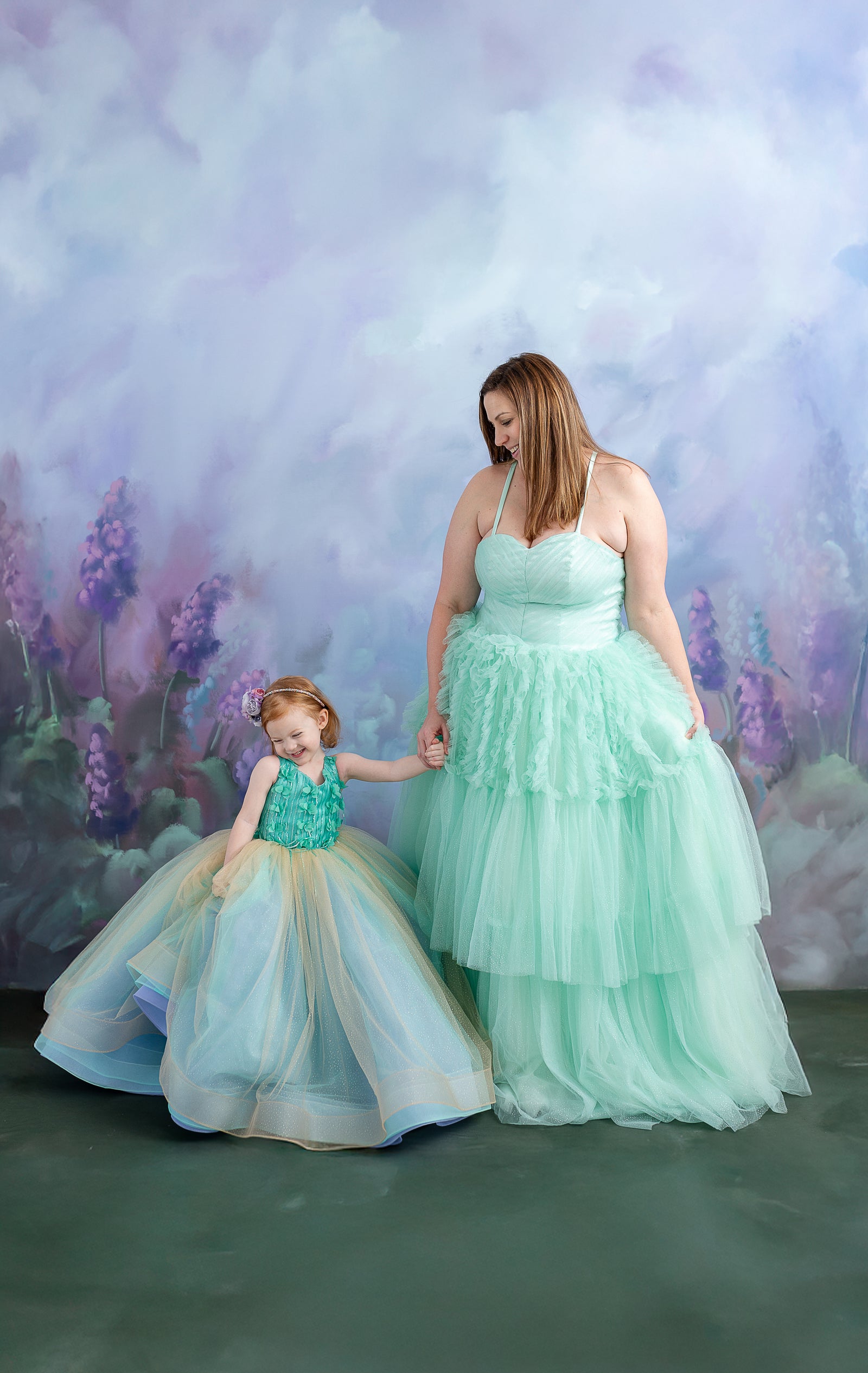A woman and child, dressed in coordinating pastel gowns, share a tender moment in front of a soft, painted backdrop.
