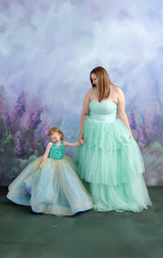 A woman and child, dressed in coordinating pastel gowns, share a tender moment in front of a soft, painted backdrop.
