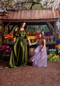 "A mother and daughter holding hands in front of a rustic flower market backdrop, wearing elegant green and lavender gowns."
