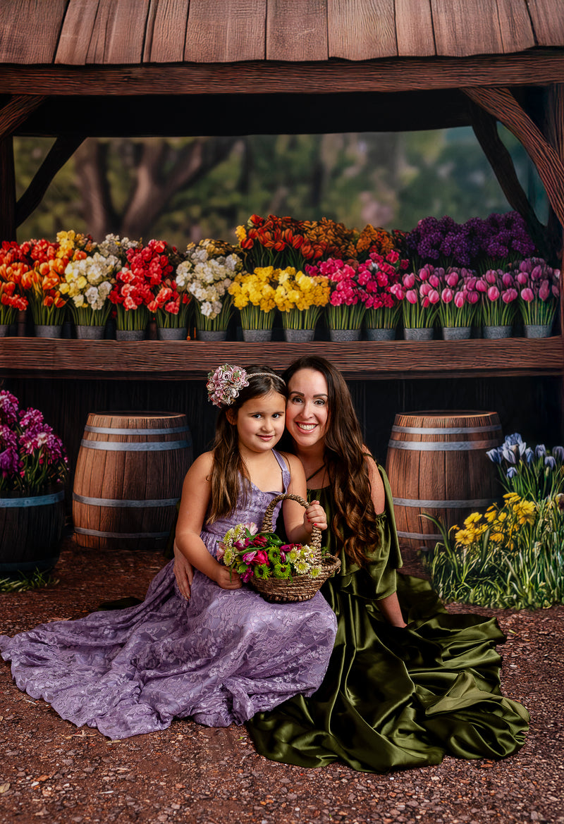 "A woman in a green satin gown smiles at a young girl in a purple lace dress, surrounded by vibrant flowers in barrels."
