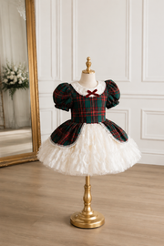 A children's dress displayed on a mannequin stand in an indoor room. The dress has a dark green and red tartan bodice with puff sleeves, a small red bow at the neckline, and a fluffy white ruffled skirt.