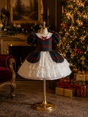 A children's holiday dress displayed on a gold dress form mannequin in a decorated living room. The dress has a dark green and red tartan bodice with puff sleeves, a white ruffled collar, and a red bow at the neckline, paired with a white ruffled skirt.