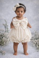 Baby in a white lace romper with a large bow standing against a floral background