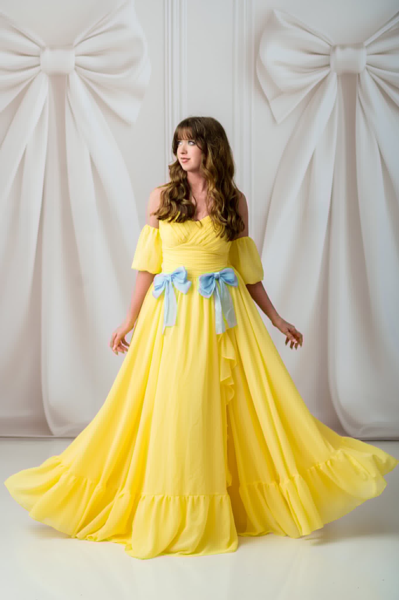 Woman in a yellow dress with blue bow details standing against a white background