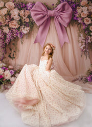 Young girl in a white dress sitting under a large pink bow with floral decorations.