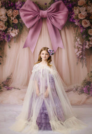 Young girl in a purple and white dress standing in front of a floral backdrop with a large pink bow.