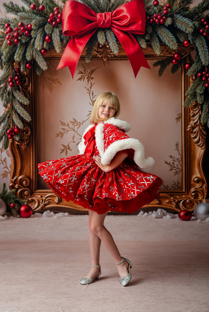 Child twirling in a red velvet sequin bow dress with puff sleeves and a lace-trimmed skirt, wearing a matching hooded cape with faux fur trim.

