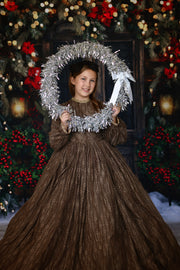 Child model in a couture floor-length lace dress with dramatic sleeves and satin tie neckline, styled for a festive Christmas scene.

