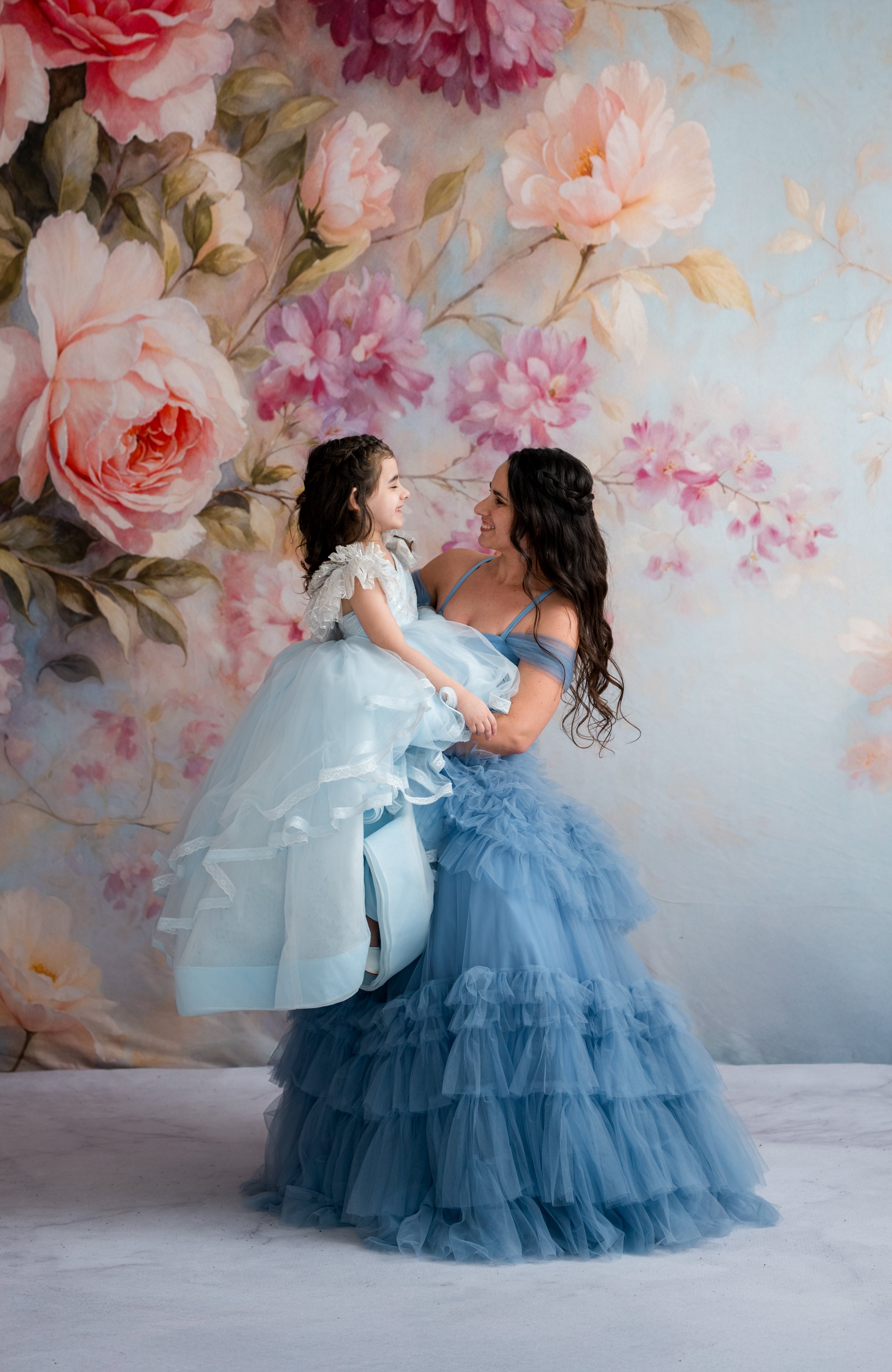 Woman in a blue dress holding a child in a white dress against a floral backdrop