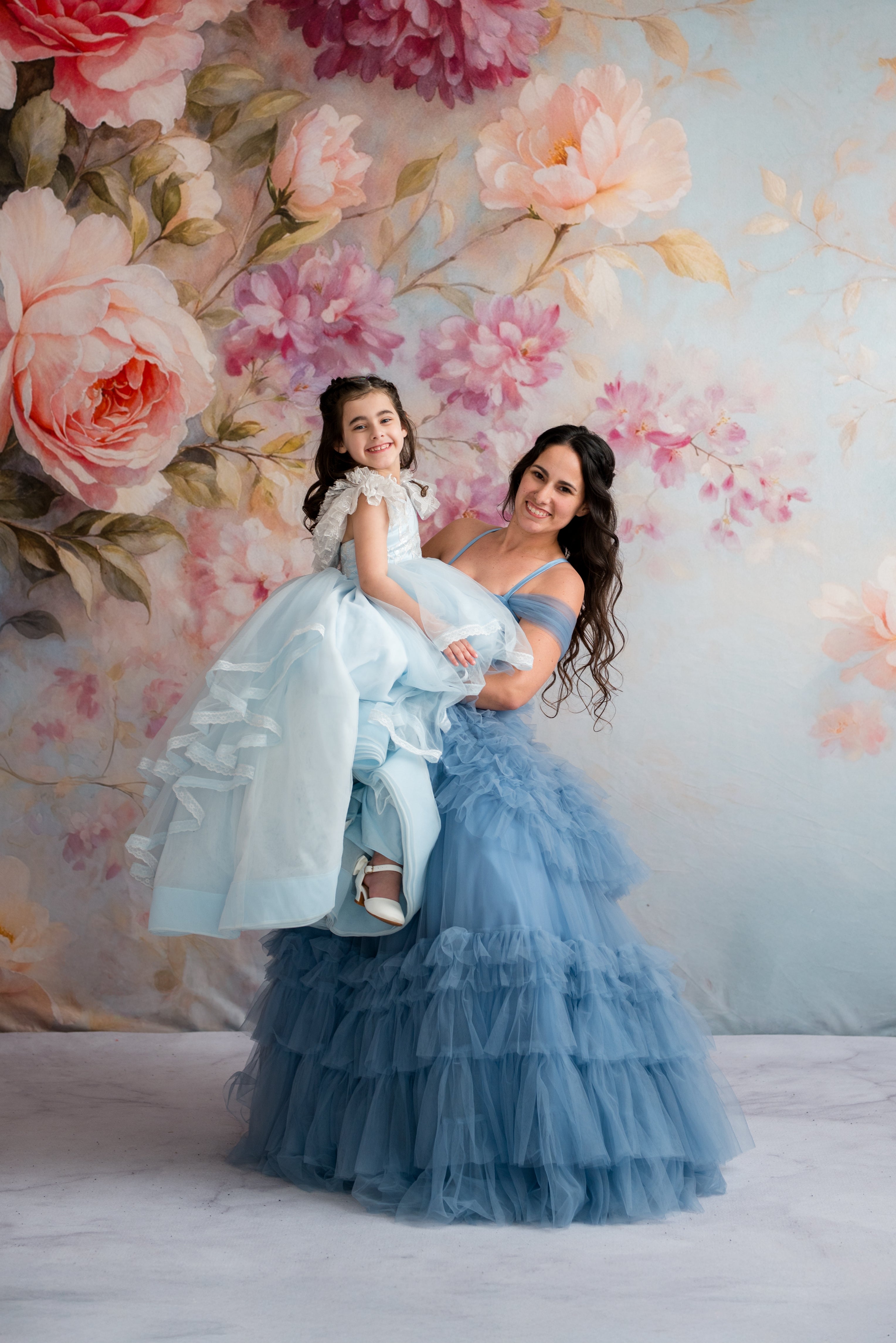 Woman in a blue dress holding a child in a light blue dress against a floral backdrop