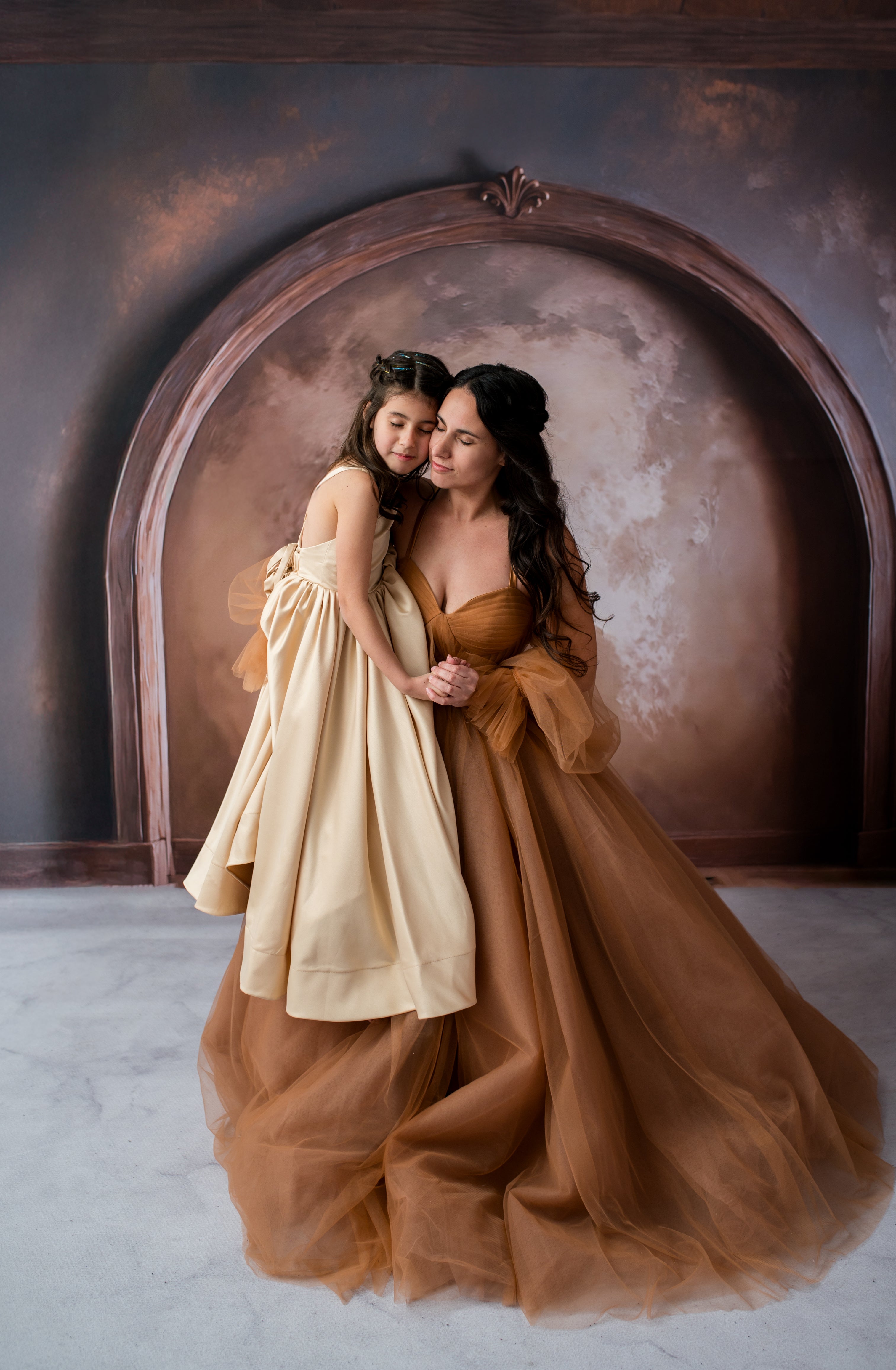 Two women in elegant dresses standing in front of a decorative arch with a moon-like background.