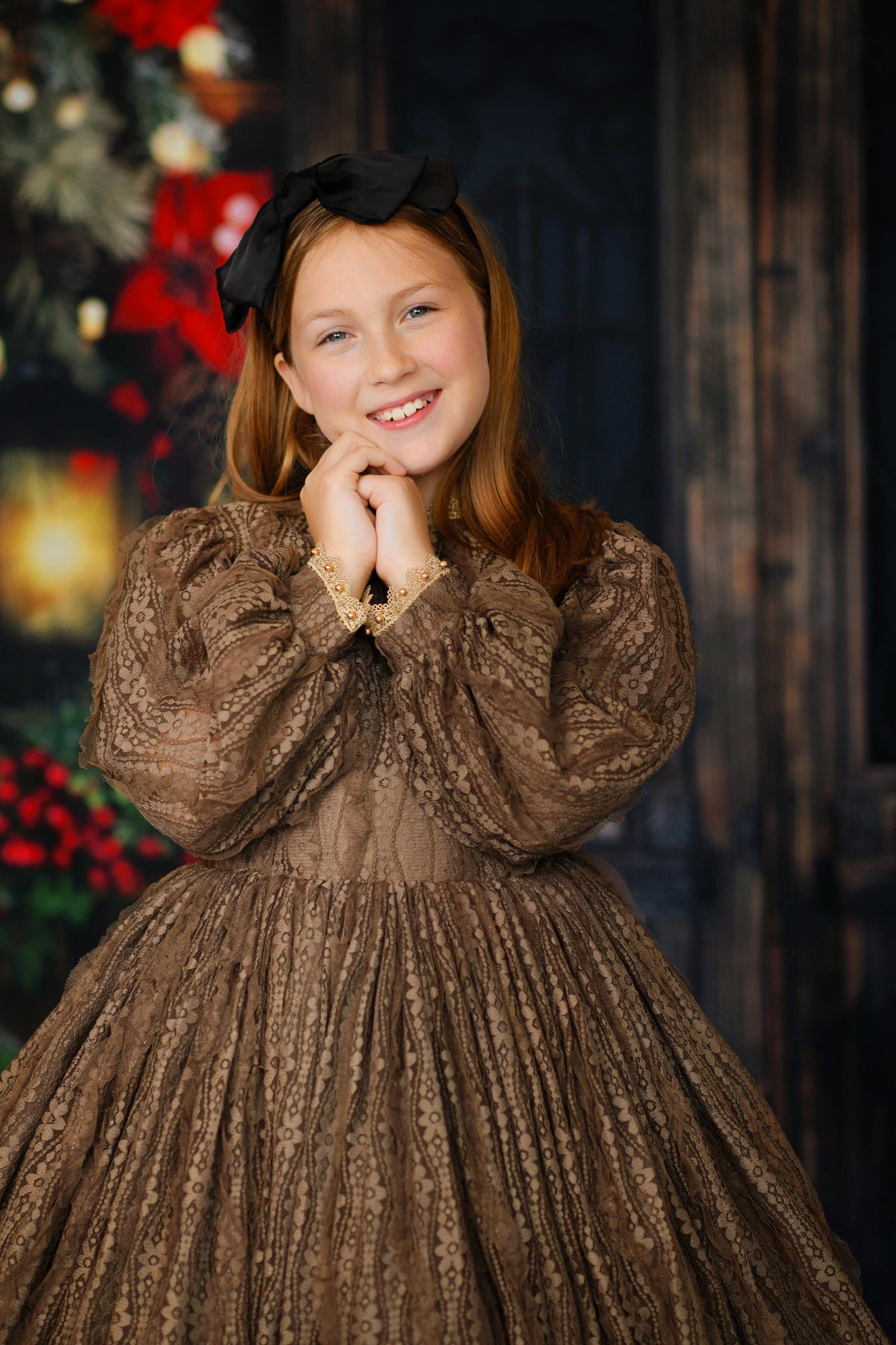 Young girl standing in front of a decorated winter door scene, wearing a brown lace gown with puff sleeves and vintage gold detailing.

