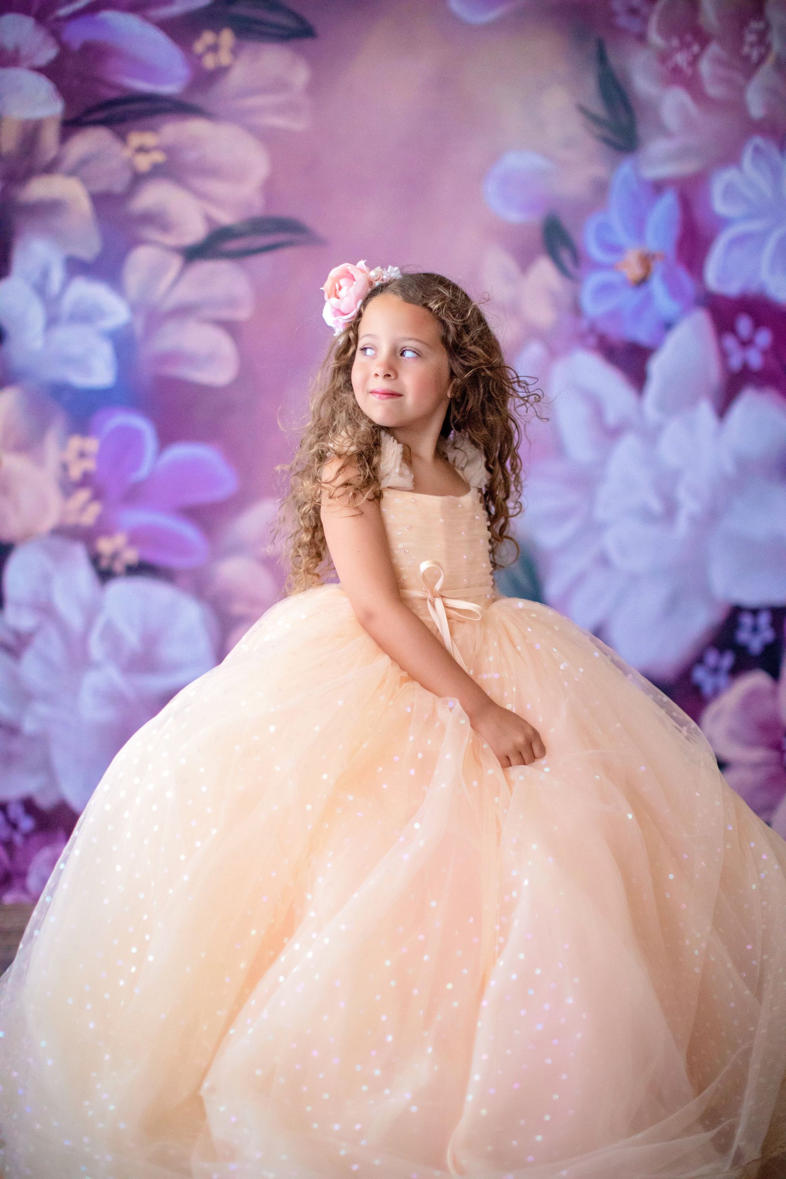 "Child wearing a full, ruffled confetti gown adorned with pastel polka dots, standing outdoors with balloons and flowers in her hair, against a scenic natural background."

