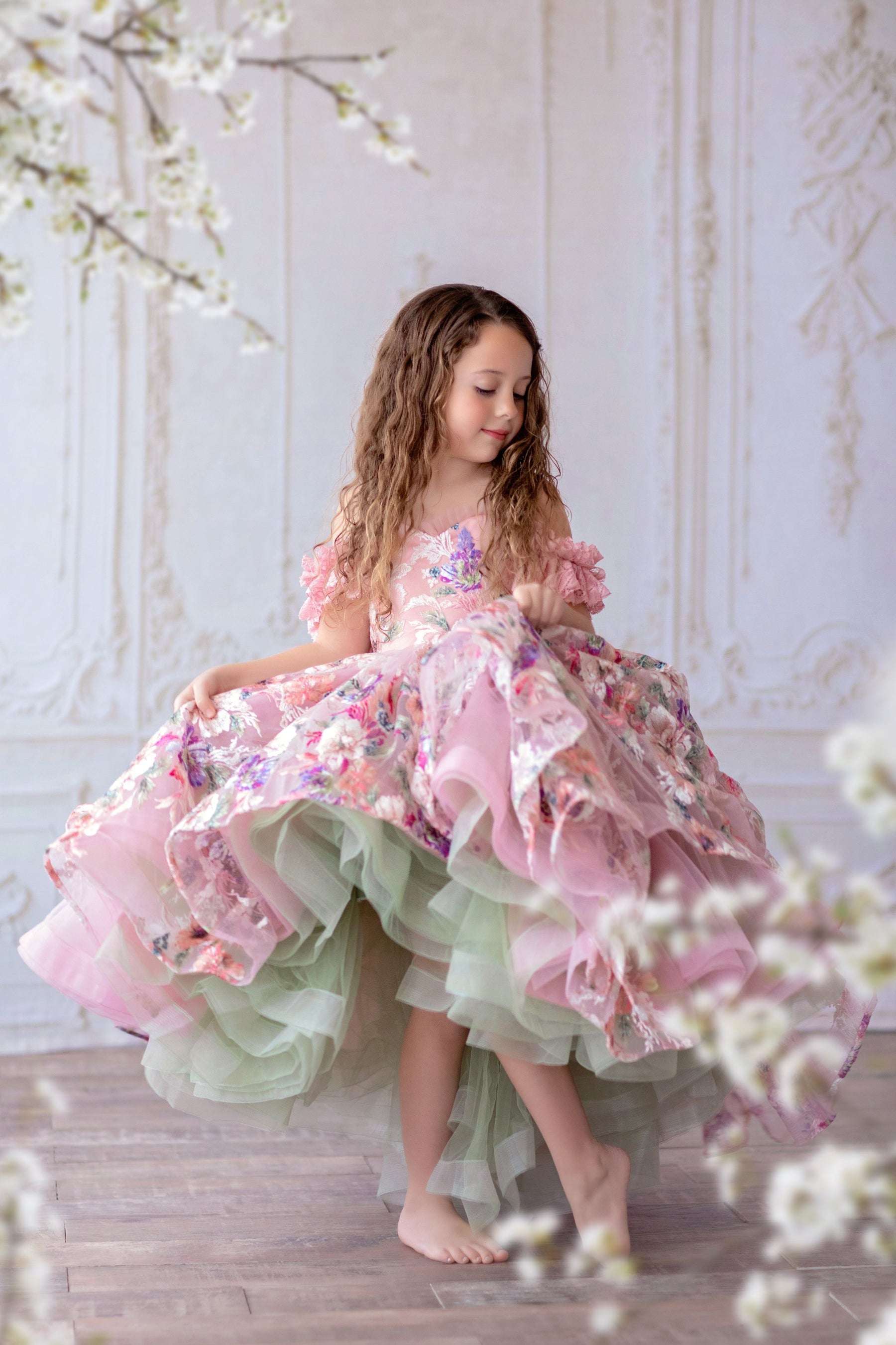 A young girl in a pink floral bouquet high-low length dress with a built-in petticoat, horsehair trim on the skirt, and a flexible back with shoulder ties.