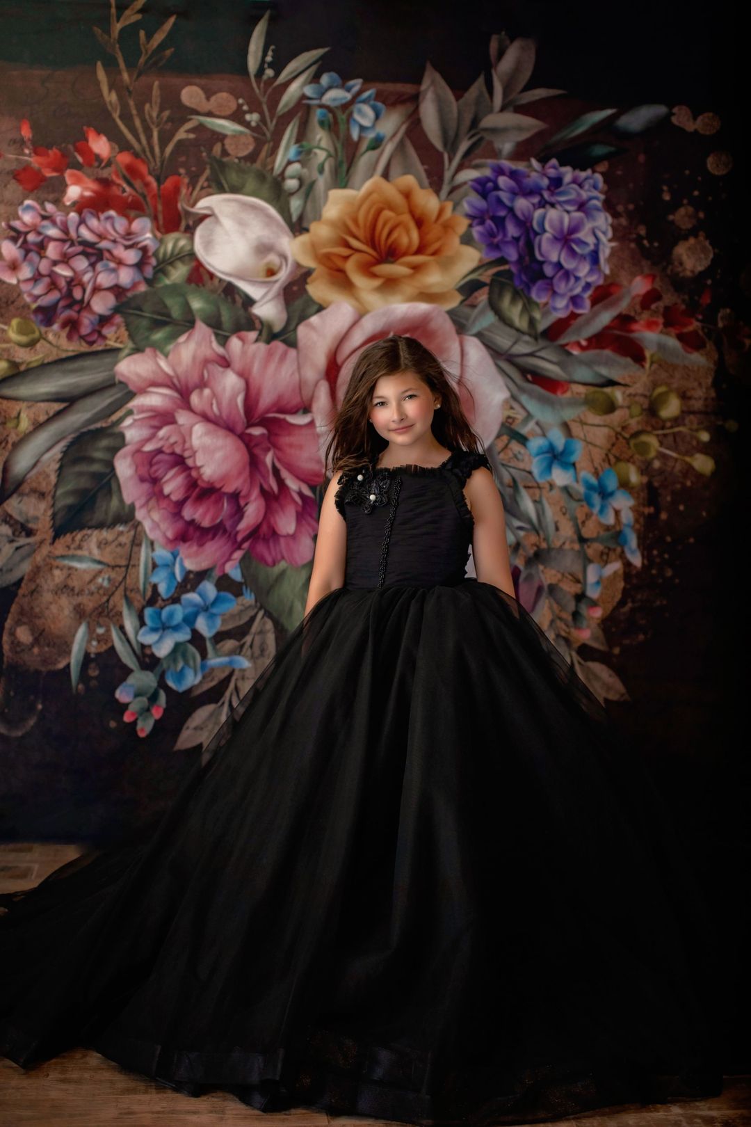 A young girl in a floral-patterned black gown with 3D butterfly details on the bodice, standing in front of a floral-themed backdrop.
